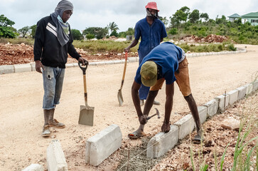 Men With Shovel Watch Their Colleague Align A Concrete Block With A Hammer. 