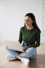 Young cute confident student woman in casual clothes is using a laptop and smiling while sitting on the floor, on gray background. Study, education, university, college, graduate concept