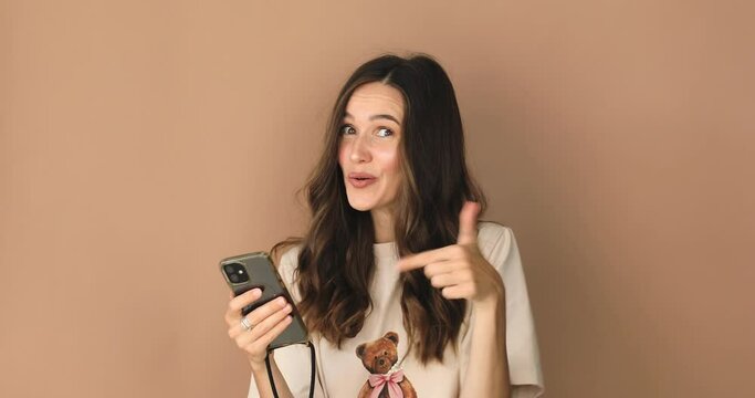 Smiling shocked happy win beautiful young woman isolated on beige background. Look surprised wow hold using mobile cell phone. Achievement, got hired, lottery money win triumph concept, thumb up.