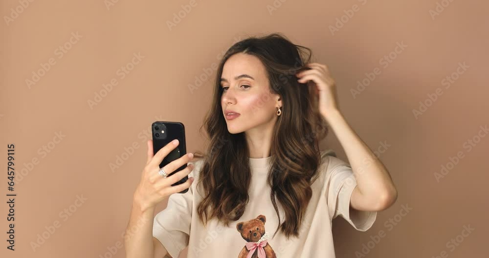 Wall mural Brunette girl stares at the phone and fixes her hair. Female uses phone like mirror. Young girl make photo from hands with phone on front mobile phone camera on beige background, look at camera.