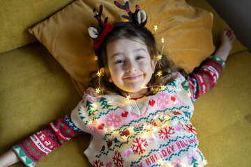 Happy cute girl in Christmas pullover playing with string light at home