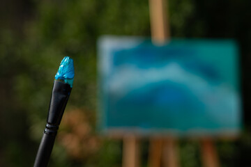 Brush with blue paint seen from up close and an easel with a contemporary art painting behind