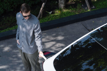 Young man travel with EV electric car charging in green sustainable city outdoor garden in summer...