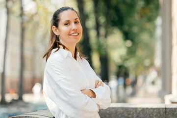Close up outdoors portrait of a wide smiling young woman with arms corssed in white shirt.