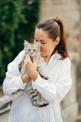 Outdoors portrait of a young woman with eyes closed embracing a grey cat on the street.