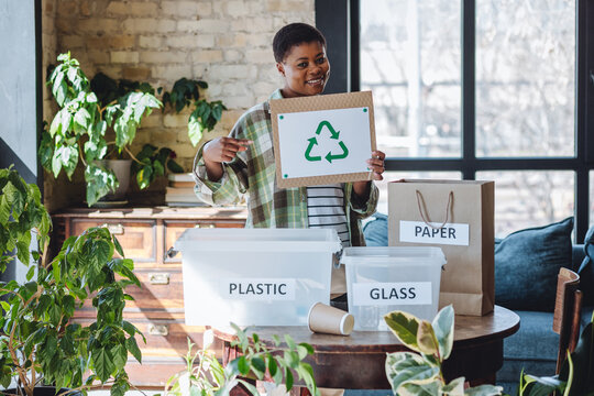 Young African American smiling female student holding recycling sign celebrating Earth Day in her house. Concept of social responsibility, sustainable lifestyle, home routine to reduce pollution.