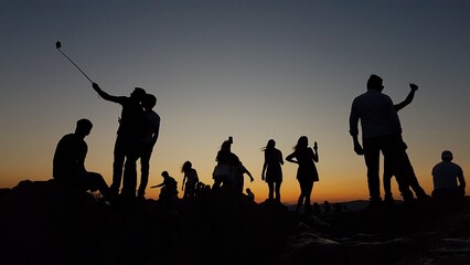 A group of diverse individuals stand on a mountain peak, taking selfies in the sunset