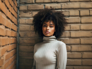A young African American woman with an Afro hairstyle, wearing a gray turtleneck, confidently posing in front of a textured stone wall.