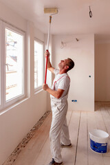 Construction worker painter man with paint roller in his hand and buckets of products to restore and paint the wall and celling, indoor the building site of a house