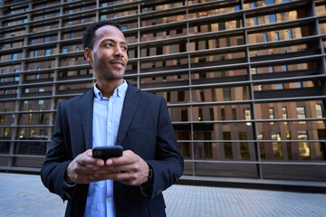 Young adult Latin professional male looking curious to side using smart phone center building...