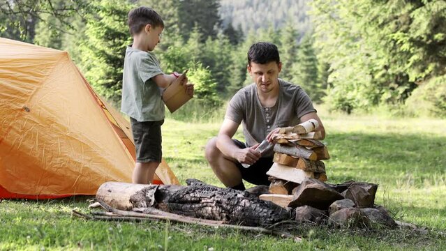 Dad and son sit together by a bundle of firewood and light a fire near the tent. The spirit of adventure and adventure. Hiking and camping. Travel concept 4K.