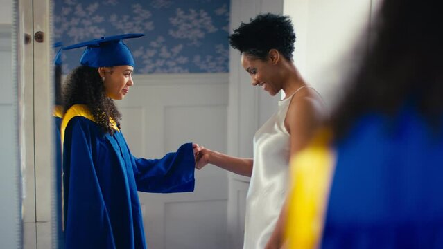 Proud mother celebrating with teenage daughter wearing graduation robes reflected in mirror at home - shot in slow motion