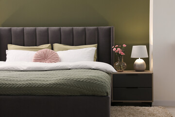 Large comfortable bed, nightstand, lamp and beautiful flowers in stylish room. Interior design