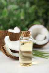 Bottle of organic coconut cooking oil, leaf and fresh fruits on white wooden table, closeup