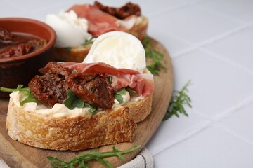 Delicious sandwiches with burrata cheese, ham and sun-dried tomatoes served on white tiled table, closeup. Space for text