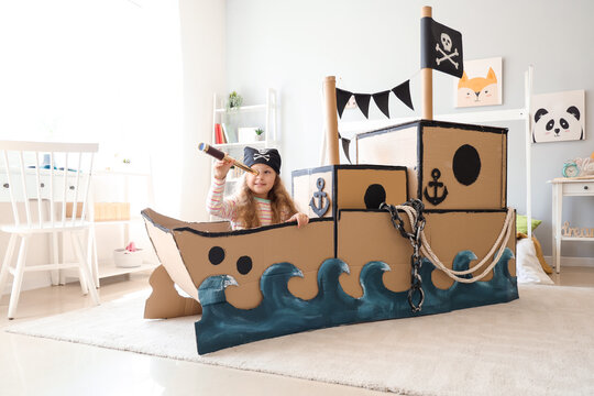 Cute little girl dressed as pirate playing with spyglass in cardboard ship at home
