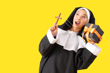Young Asian woman dressed as nun with cross and Halloween gift on yellow background