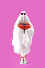 Young woman dressed as ghost with Halloween gift on pink background