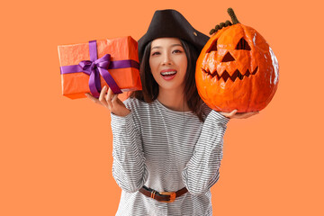 Young Asian woman dressed as pirate with Halloween pumpkin and gift on orange background