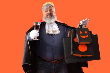 Mature man dressed for Halloween with gift bags and drink on color background