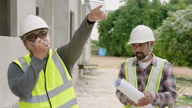 Engineer walks with blueprints to consult with Asian construction supervisor who is use walkie-talkie. Talk to crane operator. At the house construction site Wear a safety helmet and reflective vest.