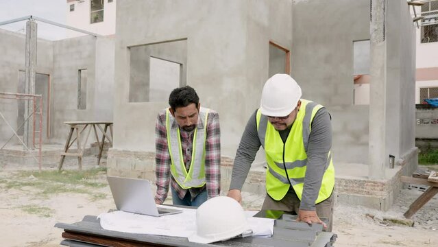 Indian engineer plans work with Asian construction supervisor. Use laptop and tablet to review house plans. With blueprints at house construction site. Man wearing a safety helmet and reflective vest