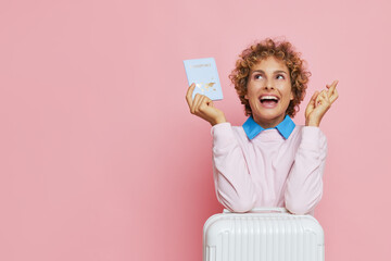 Curly-haired woman dressed in pink sweatshirt poses on pink backdrop with white suitcase and...