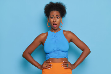 Black girl in blue top posing in blue studio with hands on her hips looking at the camera with indignation, bad mood concept, copy space