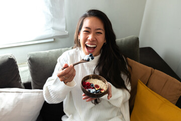 Smiling young happy Asian woman eating healthy breakfast bowl of yogurt looking at camera.