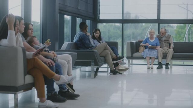 Diverse multiethnic people sit on couches in clinic lobby area, wait for appointment with doctor or medical test results. Waiting area in medical center with modern design. Healthcare system.