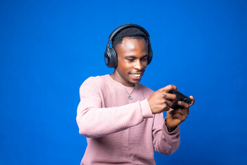 A young adult African man with a smartphone, earphones and game controller stands isolated in front of a blue colored background. 