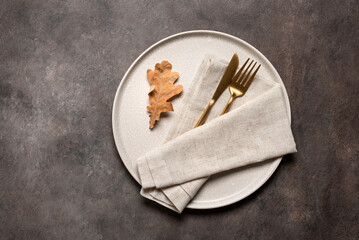Minimal autumn table setting. Beige plate, gold cutlery and beige linen napkin on a brown rustic background. Top view, flat lay, copy space.