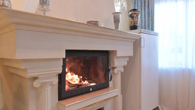 A white fireplace with a black firebox and burning flames, decorated with vases and a metal bowl. Blue curtains add color.