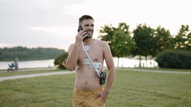 Man talks on phone, walks through park while wearing holter, monitoring heart function. Sensors attached to bare chest, record heart impulses. Concept of usual lifestyle during treatment.