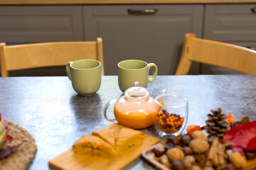 On the table are two green mugs, a teapot of sea buckthorn tea, and a pumpkin pie. In the kitchen there is a table and two empty chairs without people