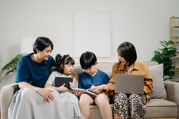 Obraz premium Asian parents and kids laugh use devices together sit on sofa, tech addicted family with children hold laptop phone digital tablet having fun at home