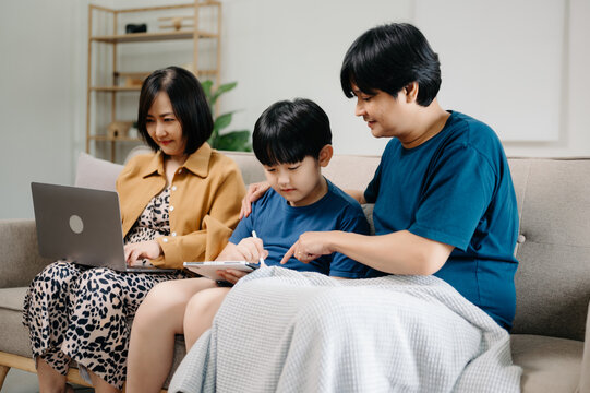 Asian parents and kids laugh use devices together sit on sofa, tech addicted family with children hold laptop phone digital tablet having fun at home