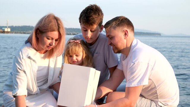 Happy family celebrating little girl's birthday on the seashore mom brings a box with cake girl opens a gift dad mom brother and sister on the ocean shore. on a sunny day