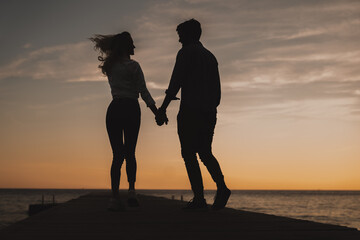 Silhouette of happy couple holding hands while standing at the sea dock. Two romantic people enjoying the sunset at beach.