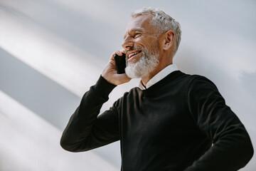 Portrait of happy senior man talking on mobile phone and smiling while standing against the wall