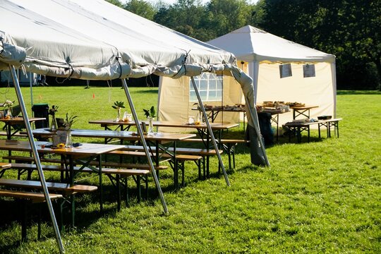 Offene Festzelte &uuml;ber Holztischen- und b&auml;nken von Sommerfest auf gr&uuml;ner Wiese vor hohen gr&uuml;nen B&auml;umen bei Sonne am Abend im Sommer
