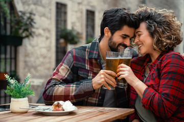 Young smiling tourist couple drinking beer in the city. Two affectionate people in love sitting in restaurant, sharing romantic moment, toasting. Curly hair woman and bearded man toasting with beer.