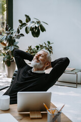 Happy mature businessman holding hands behind head and keeping eyes closed while resting in office