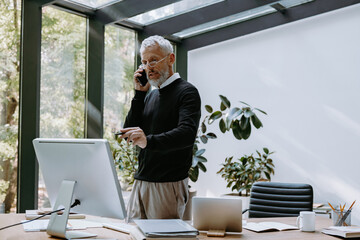 Mature man talking on mobile phone and pointing computer monitor while working in the office