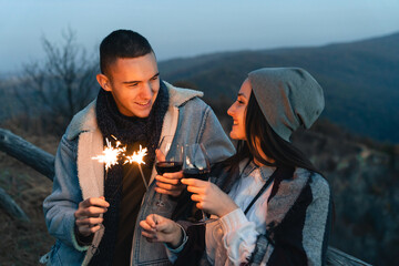Young cheerful couple lighting up a sparkling stick. Woman and man holding a glass of wine...