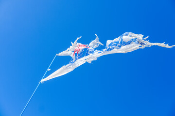 a torn flag fluttering against the blue sky, a torn plastic flag fluttering against a blue sky, a torn wind direction flag on the beach, wind direction marker on the beach, blue sky bakcground