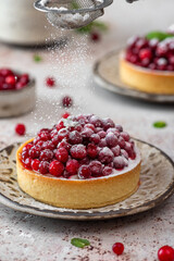 Pouring powdered sugar over tarts with fresh cowberry, cream and mint on rustic background. Berry tartlets. Vertical orientation