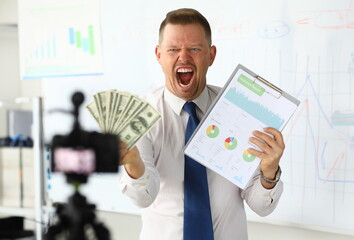 Portrait of surprised blogger holding banknotes fan and showing statistics data with graphs, charts, diagram. Guy screaming with joy at camera in office. Increase income and making money concept
