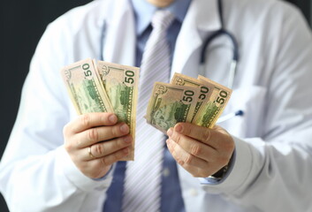 Focus on green banknotes holding by male doctor hands. Smart physician man wearing white medical uniform and stethoscope. Healthcare and clinic concept