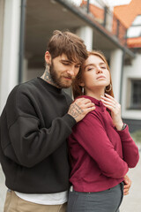 Fashionable beautiful young stylish couple in fashion hoodie on the street. Handsome hipster man with a beard hugs a beautiful red-haired woman